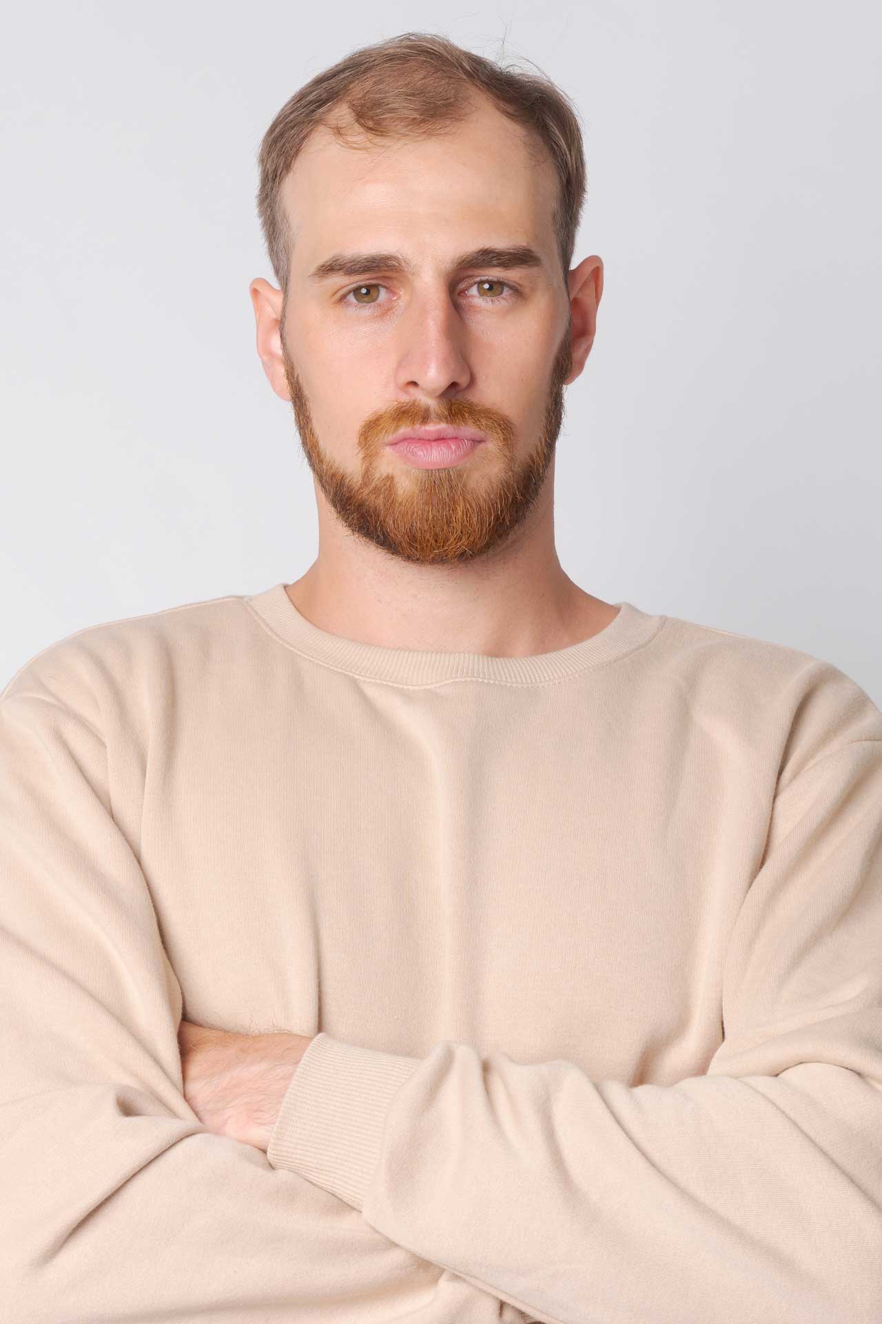 Man in a tan sweater and light brown eyes with facial hair. He is staring into the camera but not smiling with his arms crossed