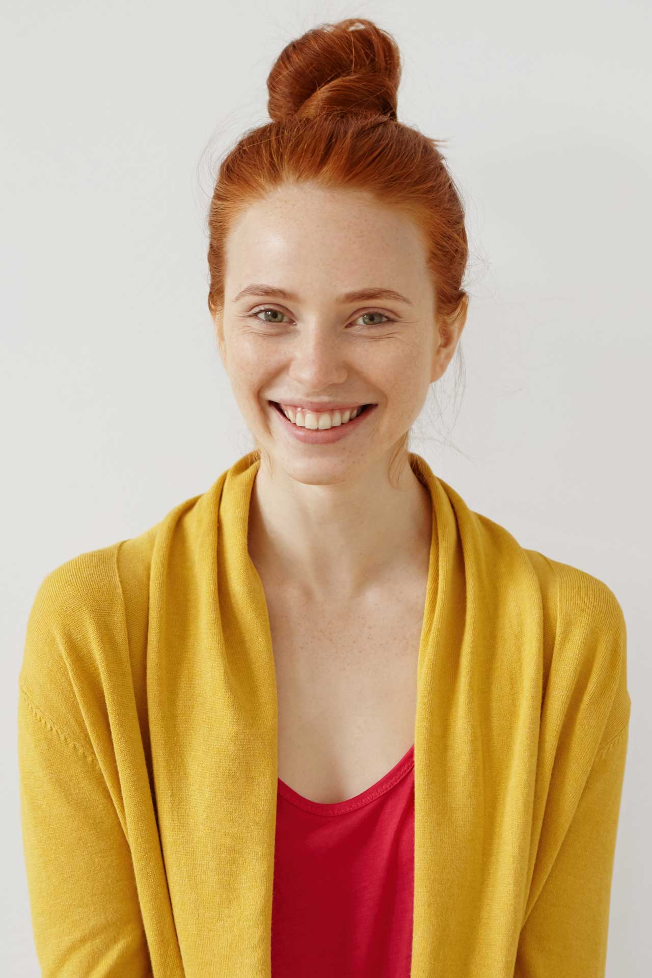 Woman with red hair smiling at the camera. She is wearing her hair in a bun and has a red shirt with a yellow sweater