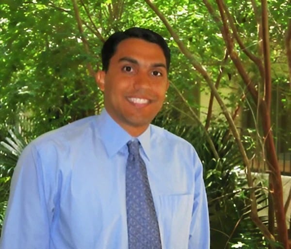 Man with dark hair smiling at the camera. He is wearing a blue shirt and tie