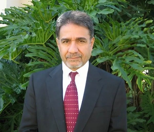 Man with dark hair, a mustache and beard, wearing a dark suit with a red tie